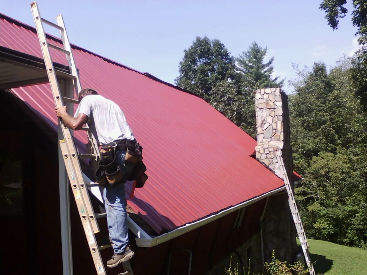 Roofer on a ladder beside a red metal roof and stone chimney — Soffit & Fascia Repair by Roofing Brothers in Tome